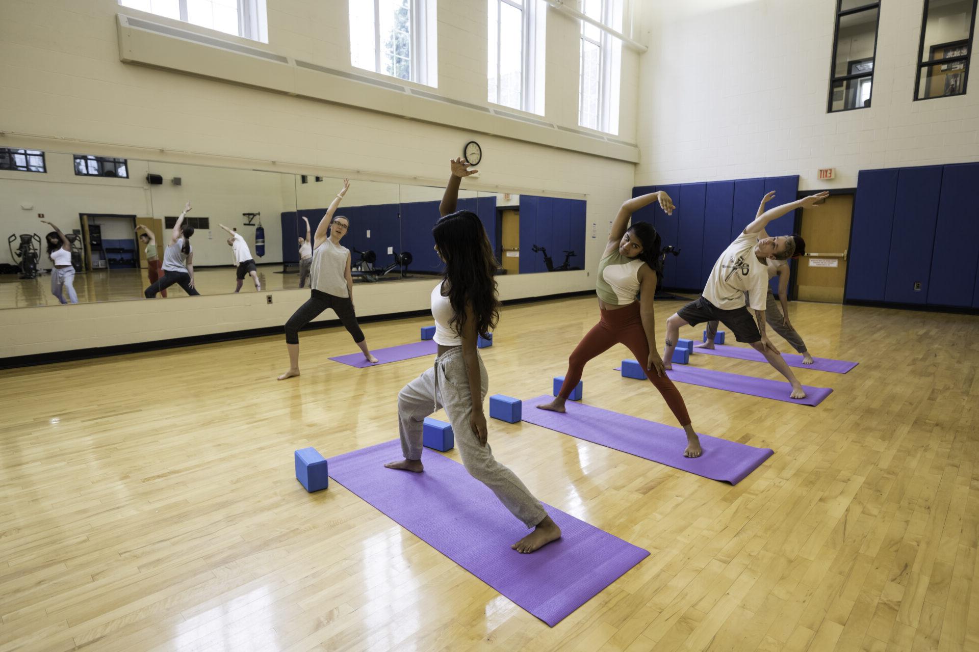 Students practicing yoga facing toward a wall with a mirror, with an instructor at the front of the room